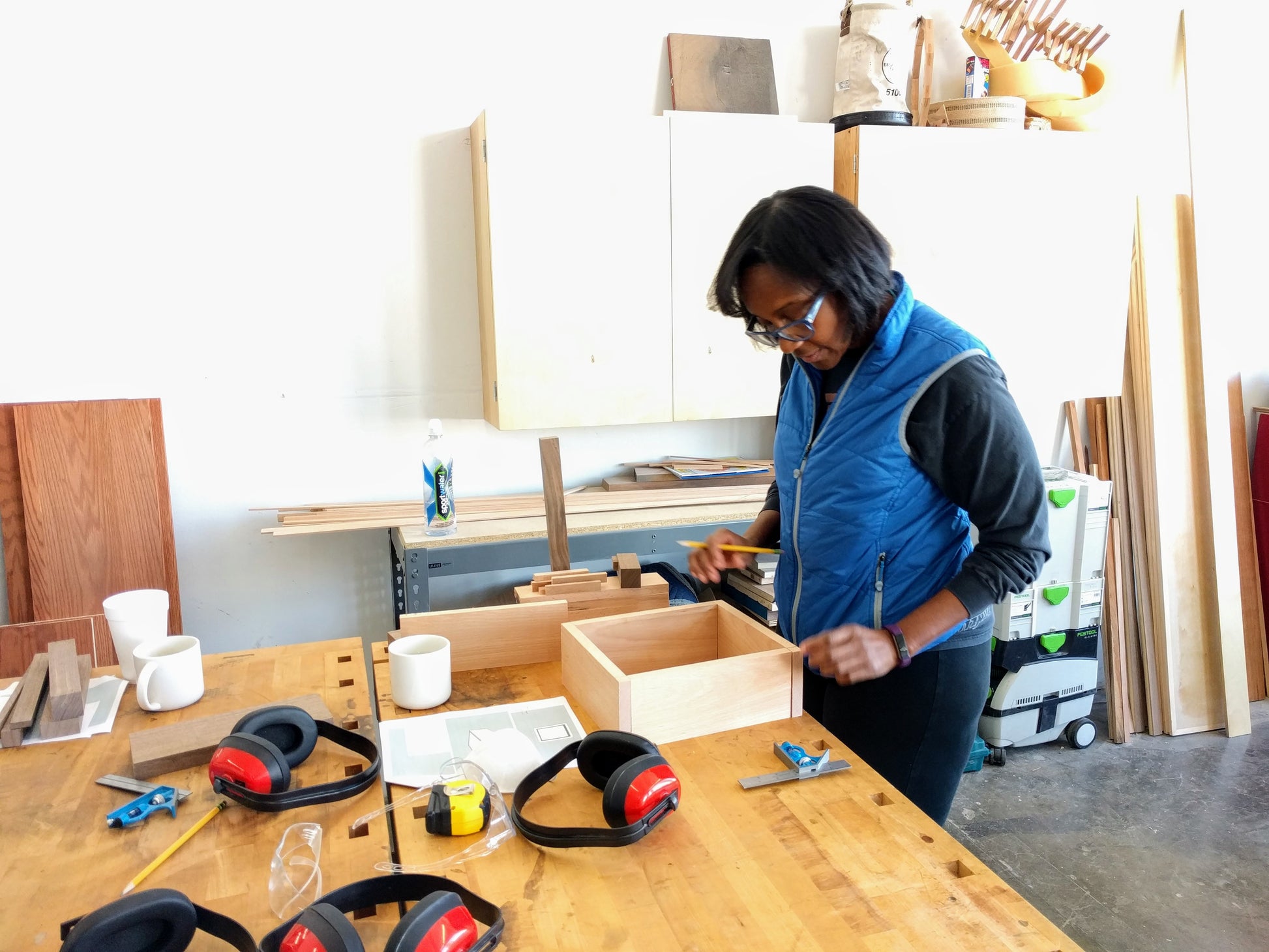 Woodworking student makes careful measurements on her box amidst strewn ear and eye protection