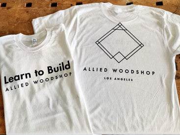 A white T-shirt on a wood surface. Learn to Build and Allied Woodshop on the front, Allied Woodshop Los Angeles and the brand logo on the back.