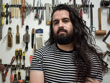 A woodworker with long dark hair and dark facial hair in a striped shirt in front of a white wall covered in woodworking hand tools.
